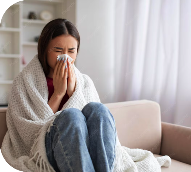 Woman wrapped in a blanket on a couch blowing her nose, looking unwell
