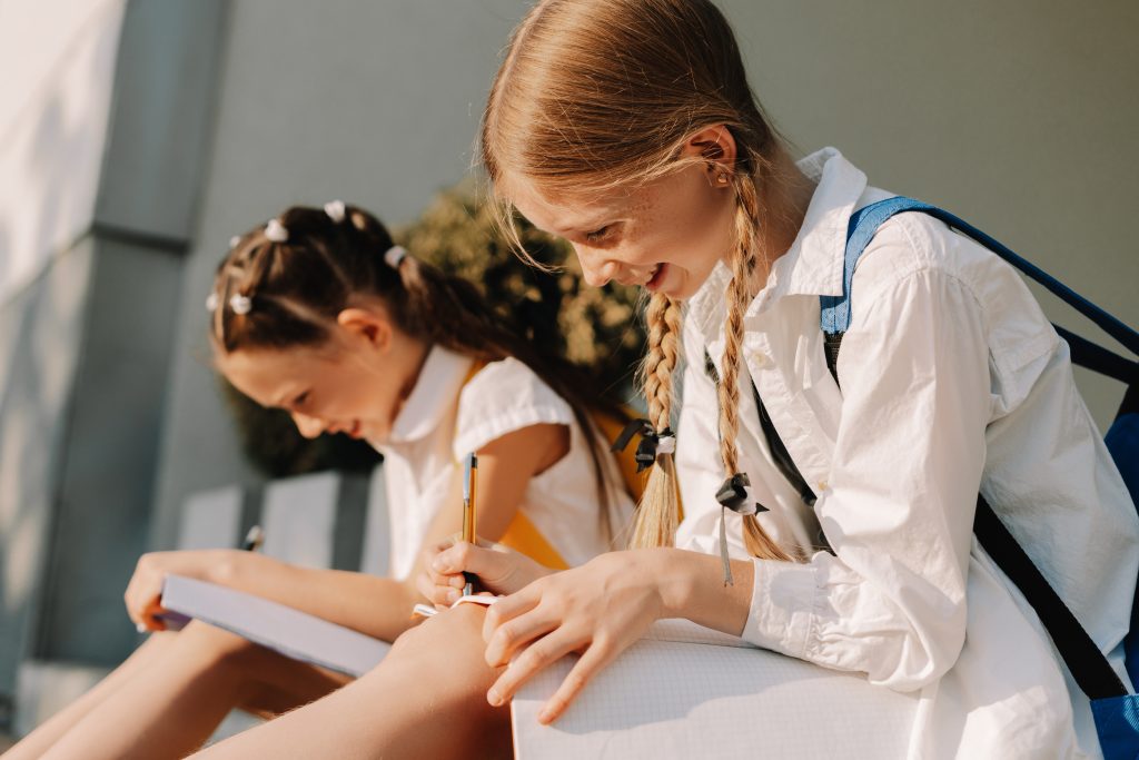 Two young schoolgirls with backpacks sitting outdoors writing in notebooks